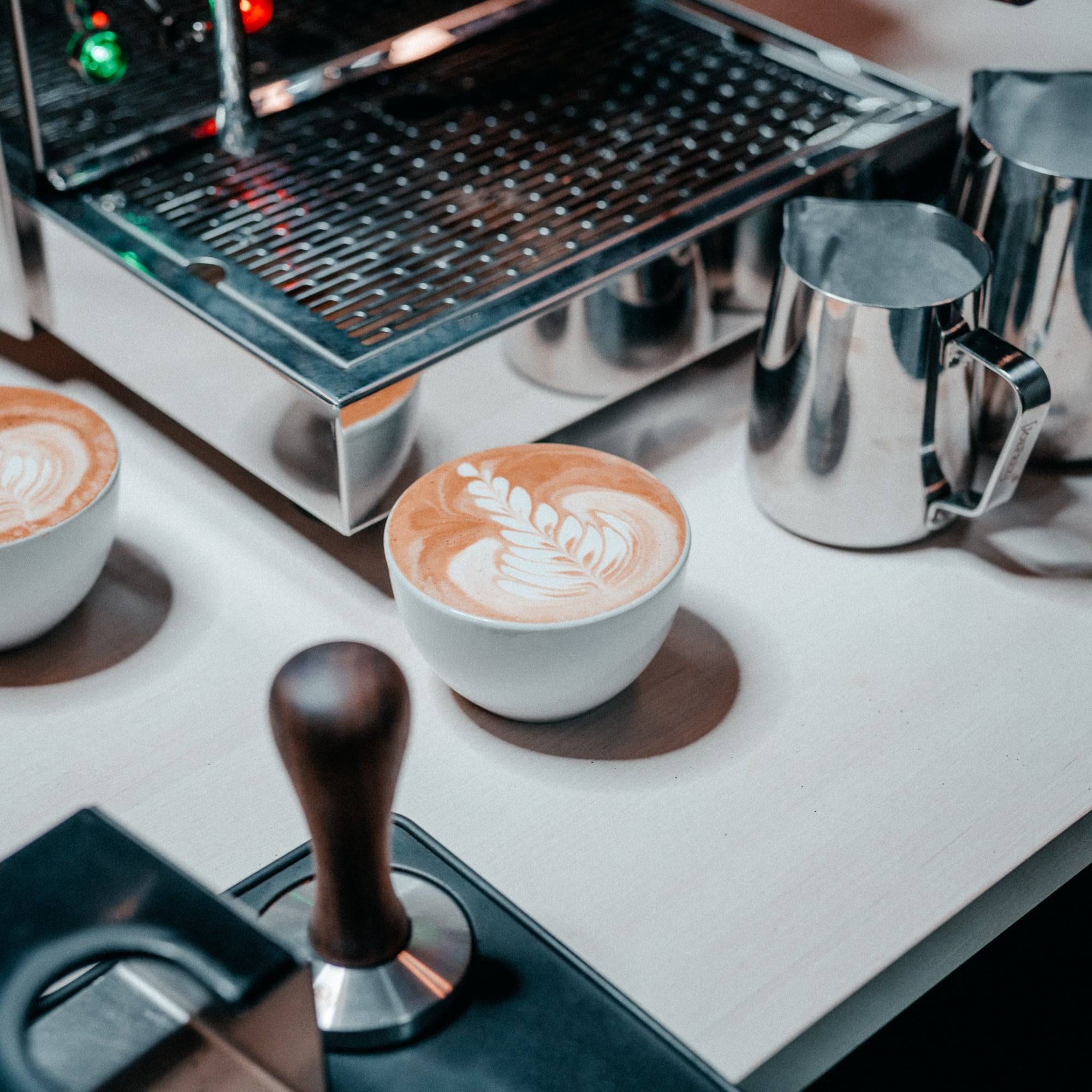 Vor einer Siebträgermaschine stehen ein Tamper, zwei Milchkannen und zwei Tassen mit Latte Art.