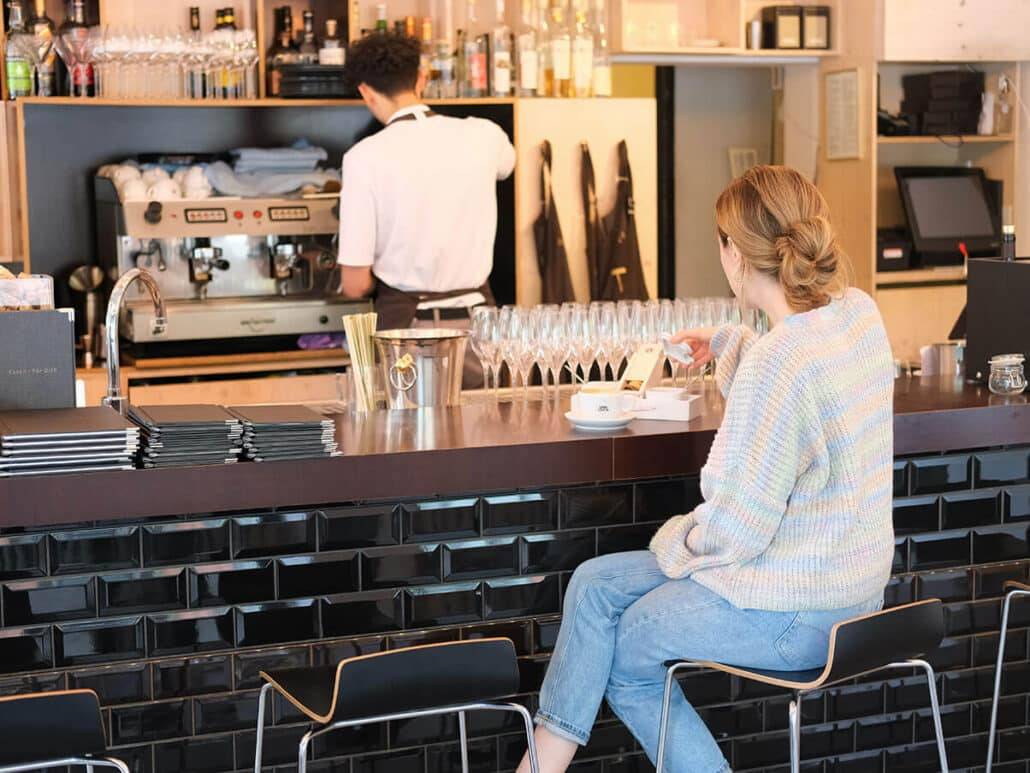 Eine Frau trinkt einen VAN DYCK Kaffee an der Theke im Restaurant Maibeck während ein Mitarbeiter einen weiteren Kaffee zubereitet.